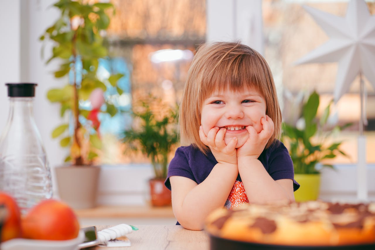 Contact A happy young girl smiling indoors surrounded by plants and a pastry, with natural light streaming in.