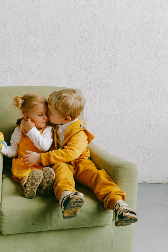Services Little brother and sister in casual wear sitting in cozy armchair together and hugging in living room