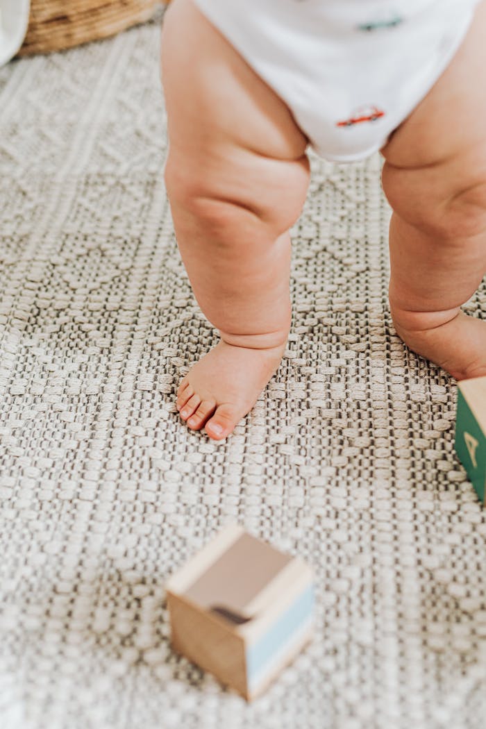 Home Candid shot of a baby's legs standing on carpet with toy blocks scattered around.
